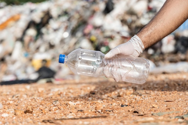 Hand, die eine Plastikflasche vor einem großen Berg und einem Müllhaufen hält. Abfallkunststoffe aus Industrie und Kommune. Weltumwelttag.