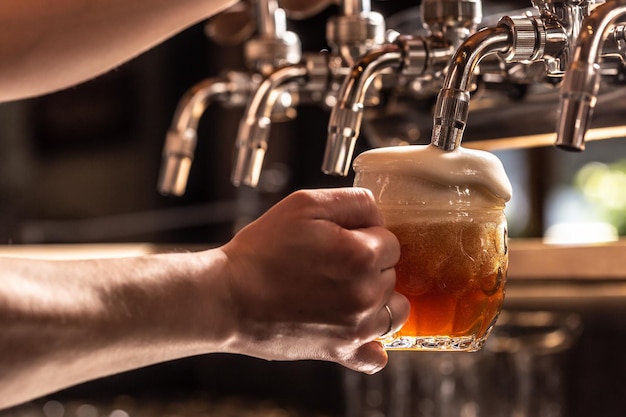 Hand, die einen Bierkrug hält, der aus dem Wasserhahn gefüllt wird, wobei Schaum aus dem Glas schwimmt