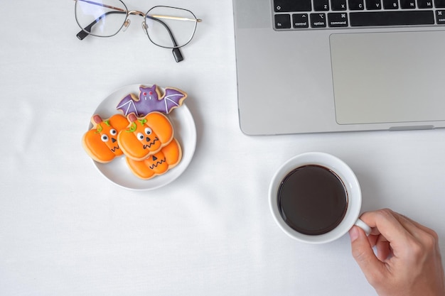 Hand, die Kaffeetasse hält und Halloween-Plätzchen während der Verwendung des Computer-Laptops isst Fröhliches Halloween-Online-Shopping Hallo Oktober Herbst Herbst Festliches Party- und Urlaubskonzept