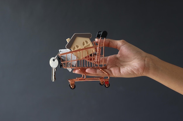 Hand, die Miniatureinkaufswagen mit vorbildlichem Haus und Hausschlüsseln gegen grauen Hintergrund hält.