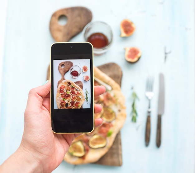 Hand, die Schuss am Handy macht: rustikale selbst gemachte Pizza mit Feigen-, Prosciutto- und Mozzarellakäse, dunkles hölzernes Umhüllungsbrett über hellblauem Hintergrund. Ansicht von oben.