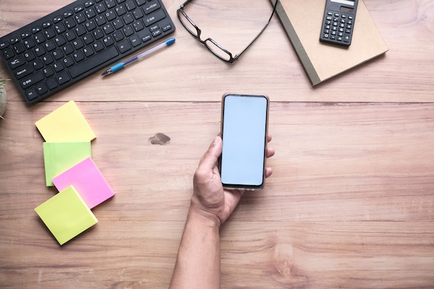 Hand, die Smartphone auf Schreibtisch mit Bürolieferanten hält