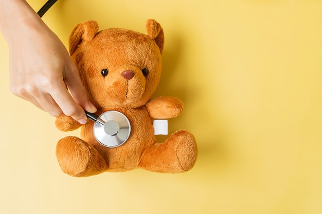 Hand, die Stethoskop auf Bärenpuppe hält, um das Leben und die Krankheit von Kindern zu unterstützen. September Aufklärungsmonat für Kinderkrebs, Gesundheits- und Lebensversicherungskonzept