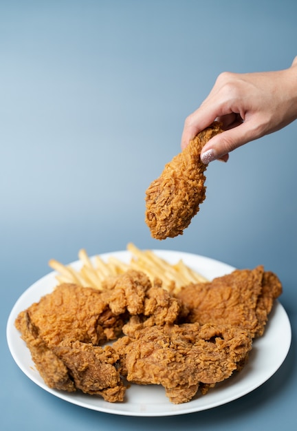 Hand, die Trommelstöcke, knuspriges gebratenes Huhn mit Pommes Frites im weißen Teller auf blauem Hintergrund hält.