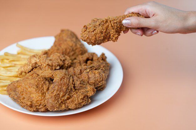 Hand, die Trommelstöcke, knuspriges gebratenes Huhn mit Pommes Frites in der weißen Platte auf orange Hintergrund hält.