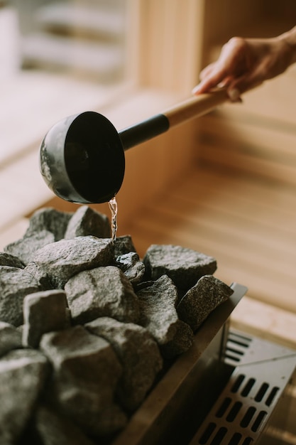 Hand einer jungen Frau, die Wasser auf heiße Felsen in der Sauna gießt