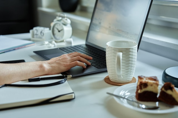 Hand einer nicht erkennbaren person, die einen laptop am arbeitsplatz in der nähe des fensters benutzt, bequeme fernarbeit zu hause