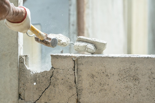 Hand eines Arbeiters, der mit einem Hammer an einer Ziegelsteinmauer auf einer Baustelle zerbricht und zerstört