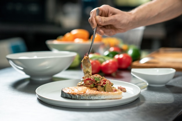 Hand eines jungen männlichen Kochs, der gebratenes Gemüse auf ein Stück gebratenen Lachs auf den Teller legt, während er leckeres Essen für den Kunden des Restaurants zubereitet