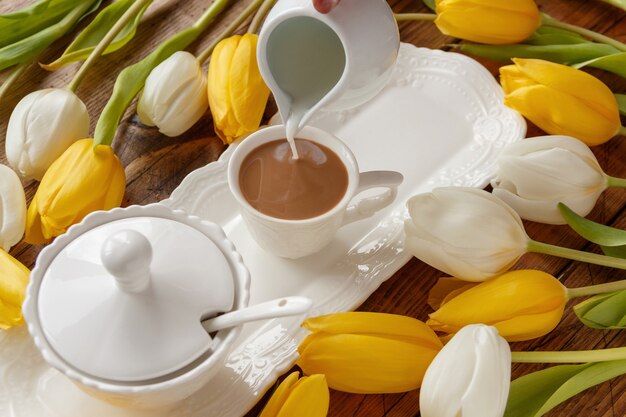 Hand gießt Milch in Kaffee in der Nähe von weißen und gelben Tulpen auf Holztisch