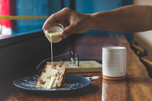 Hand gießt Vanillepuddingcreme in Obstkuchen mit Tasse Kaffee auf Tisch.