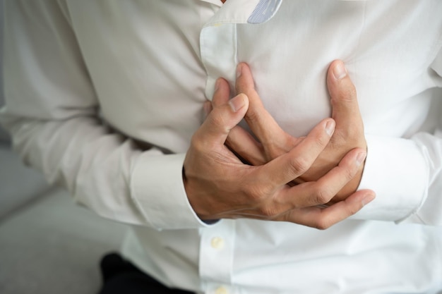 Hand hält die Brust mit Symptomen eines Herzinfarkts. Ein asiatischer Mann, der hart arbeitet, hat Schmerzen in der Brust, die durch eine Herzerkrankung verursacht werden, und eine Leckerweiterung. Das vergrößerte Herzkranzgefäß drückt mit einem schmerzhaften Gesichtsausdruck auf die Brust