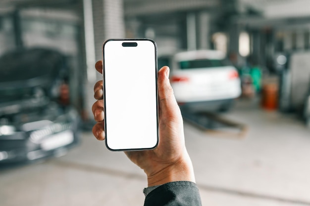 Hand hält ein Smartphone mit einem leeren weißen Bildschirm in einer Autoreparaturwerkstatt mit verschwommenen Fahrzeugen und Garagengeräten im Hintergrund