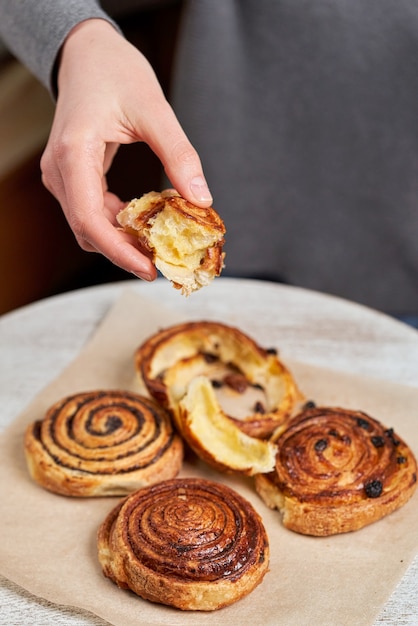 Hand hält ein Stück Butterrolle über den Tisch mit Brötchen mit Mohn und Rosinen. Vertikaler Schuss