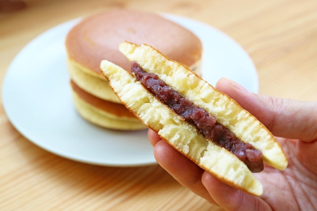 Hand hält ein Stück halbes Dorayaki mit verschwommenem Stapel ganzen Kuchens auf einem Holztisch