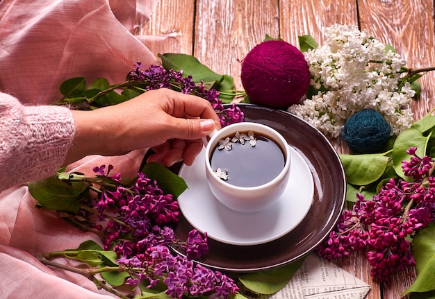 Hand hält eine Tasse Morgenkaffee mit blühenden Fliederblumenzweigen des Frühlings