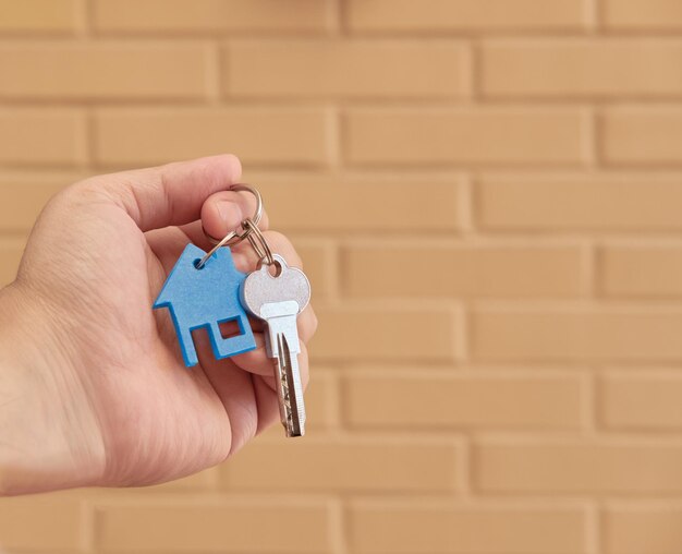 Hand hält einen Schlüsselring mit einem Schlüssel und ein kleines blaues Haus über einem Backsteinhintergrund. Real State