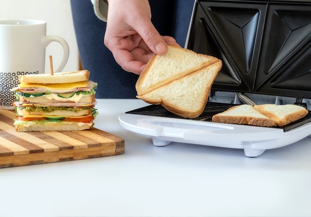 Hand hält gebratene Brotscheibe aus dem Toster in der Nähe von gekochtem frischem Sandwich mit Käse, Speck und Gemüse.