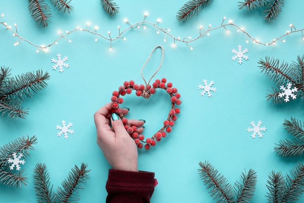Hand hält Herzkranz mit gefrosteten Beeren Winter Weihnachten flach Hintergrund mit Tannenzweigen Schneeflocken Girlande mit Weihnachtslichtern Monochromatischer blauer Minzhintergrund