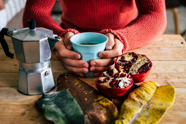 Hand hält Kaffeetasse mit halbierten Granatapfel auf dem Tisch im Restaurant. Nahaufnahme von Händen, die während des Frühstücks im Café eine warme Kaffeetasse mit frischem Obst auf einem Holztisch halten.