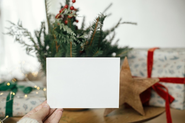 Hand hält leere Grußkarte an einem stilvoll verpackten Weihnachtsgeschenk, rustikaler Stern und Tannenzweige mit Lichtern auf einem Holztisch. Weihnachtskarten-Attrappe. Platz für Text. Postkarte mit Weihnachtsgrüßen