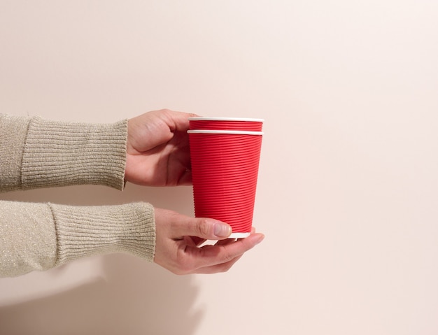 Hand hält rote Pappbecher für Kaffee, beige Hintergrund. Umweltfreundliches Geschirr, null Abfall