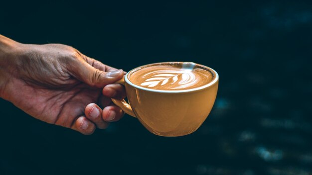 Hand Hold Cup Hot Latte Art in gelbem Becher vor dunklem Hintergrund.