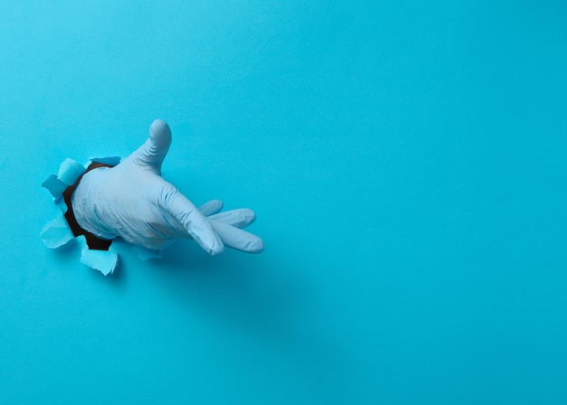 Hand in einem blauen medizinischen Handschuh ragt aus einem zerrissenen Loch in einem blauen Papierhintergrund heraus