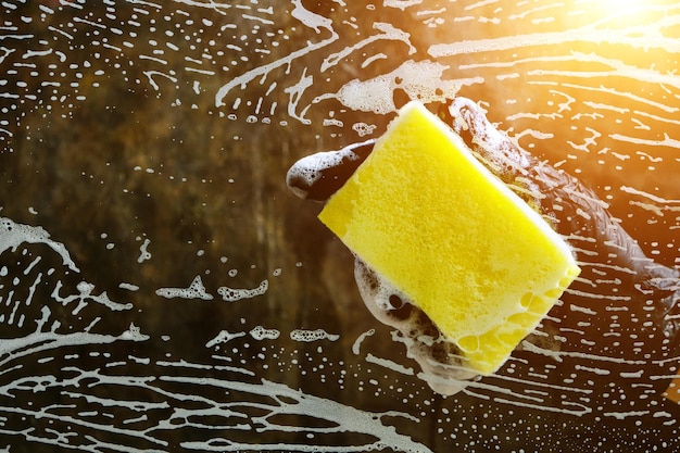 Hand in einem schwarzen Handschuh wäscht das Glas mit einem Schaumschwamm mit Schaum auf dunklem Hintergrund mit Licht.