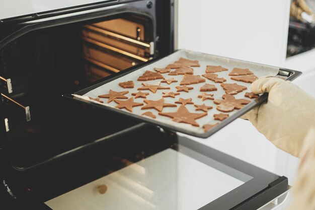 Hand in Ofenhandschuhen, Tablett mit Pfefferkuchen in weihnachtlichen festlichen Formen in modernem Ofen in weißer Küche, Weihnachtspfeferkuchen machen