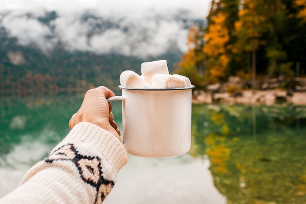 Foto hand mit becher kakao, marshmallow auf dem seehintergrund in den bayerischen alpen, deutschland