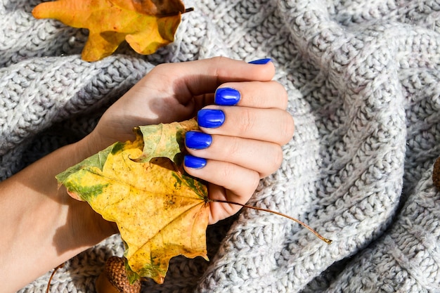 Hand mit blauen Nägeln auf gemütlichem Hintergrund des Pulloverherbstes. Weibliche Maniküre. glamouröse schöne Maniküre. Winter- oder Herbstmaniküre in Blau. Modische Nagelgellackfarbe