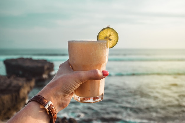 Hand mit Glas Fruchtcocktail Ozeanwellen Steine und Strand Sonnenuntergang an