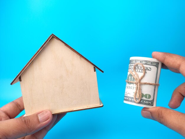 Hand mit Holzhaus und Banknoten auf blauem Hintergrund Geschäftskonzept