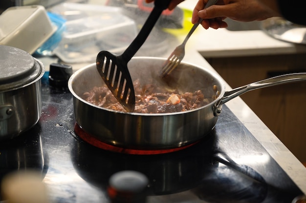 Hand mit Küchenflosse, die geschnittenes Rindfleisch in der Pfanne auf Elektroherd braten