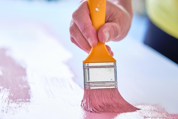 Hand mit orangefarbenem Pinsel im Vordergrund, der ein rosafarbenes Holzbrett malt