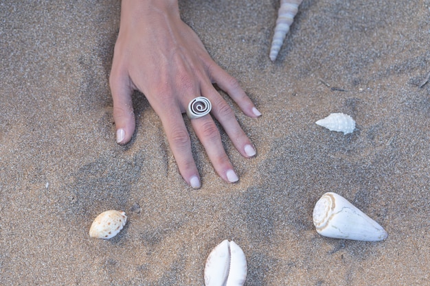 Hand mit Ring auf dem Sand an einem Strand mit Muscheln herum