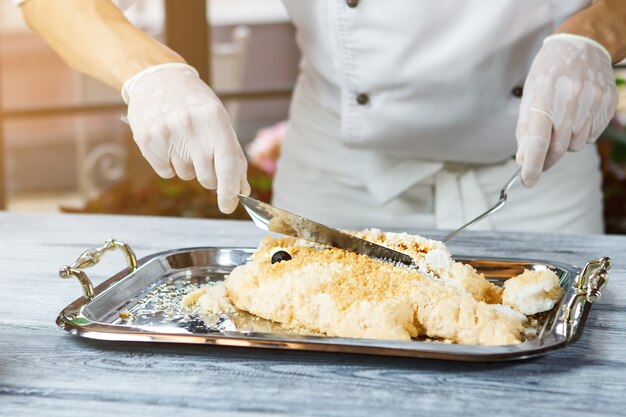 Hand mit Spachtel berührt Fisch. Tablett mit paniertem Fisch. Gekochter Dorado-Fisch in Panade. Koch, der mit Meeresfrüchten arbeitet.