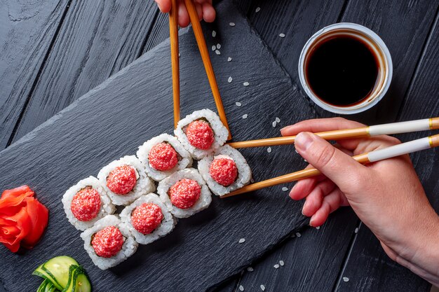 Hand mit Stäbchen nimmt Sushi von Seth-Sushi-Rollen mit Frischkäse, Reis und Lachs auf eine schwarze Tafel, die mit Ingwer und Wassabi auf einem dunklen Holztisch dekoriert ist. japanisches Essen