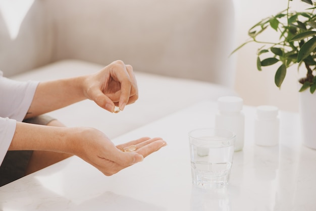 Hand nimmt gelbe Kapseln von Omega 3, weiße Pillen von Glucosamin und Kalzium auf Plastikbox, Glas mit Wasser am Holztisch, Draufsicht.