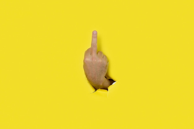 Foto hand of man making insulting gesture with middle finger through a hole in yellow paper background