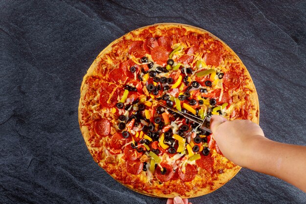Hand schneidet die höchste Pizza mit Tomaten, Salami, Oliven auf schwarzem Tisch
