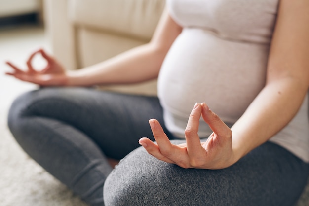 Hand und Bein im Knie einer jungen schwangeren Frau in Aktivkleidung gebeugt, die in der Haltung des Lotus sitzt und zu Hause auf dem Boden meditiert