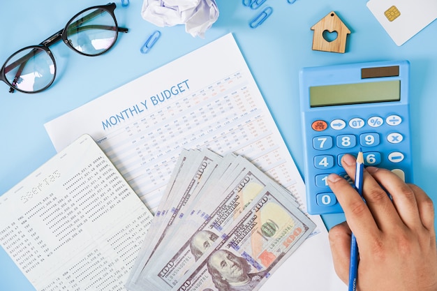 Hand unter Verwendung des Taschenrechners, der Monatsbudget mit Sparbuch und uns Banknotenebene berechnet, lag auf blauem Hintergrund