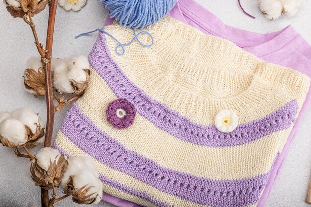 Handgefertigtes gehäkeltes Baby-T-Shirt in Fliedertönen. Material und Requisiten enthalten Garnhaken, Stricknadeln und Basteldekor. Heller Steinbetonhintergrund, Draufsicht