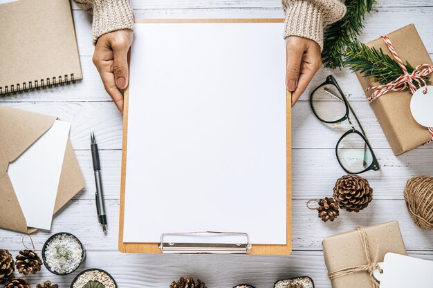 Handgehaltenes Clipboard auf einem weißen Holztisch mit Brille, Stift und getrockneten Kiefernkegel