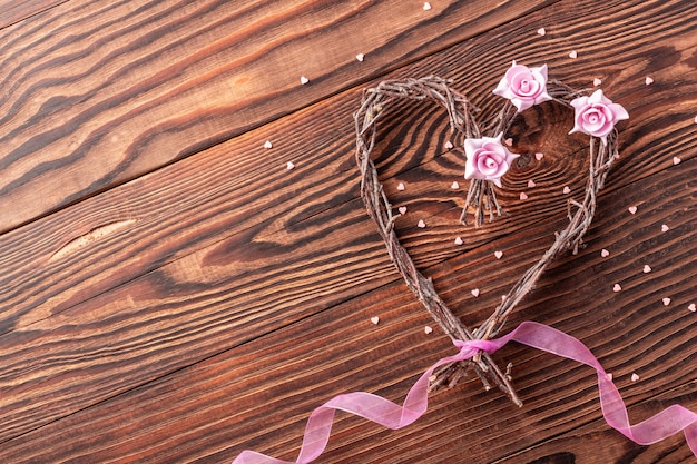 Handgemachtes Zweigherz mit Blumen und rosa Band auf Holzhintergrund für Hochzeit des Valentinstags