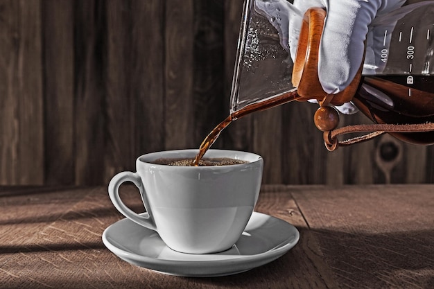 Handgetragen in weißem Handschuh gießt frisch gebrühten, leckeren Kaffee aus dem Kaffeetropfer in eine Keramiktasse
