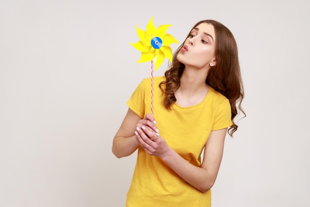 Handmühle Porträt eines glücklichen, braunhaarigen Teenager-Mädchens im urbanen Stil, gelbes T-Shirt, das auf Papierwindmühlenwindrad-Spielzeug auf Stick bläst. Studioaufnahme isoliert auf grauem Hintergrund