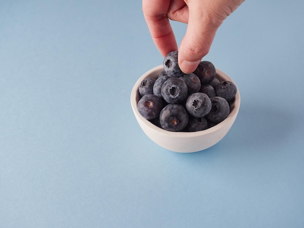 Handpflücken einer Blaubeere aus einer kleinen Schüssel auf blauer Oberfläche mit Kopierraum